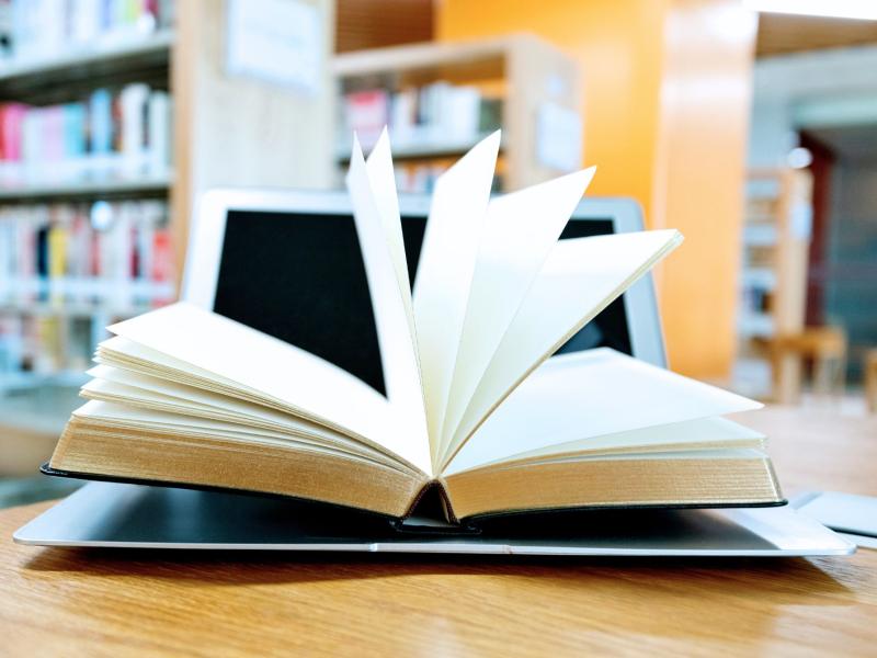 Opslået bog med sider i bevægelse foran en computer på et bibliotek.